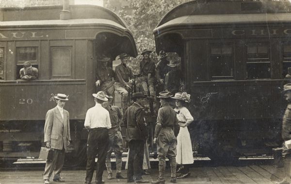 A group of men and women at a train station near two railroad cars. Several stand on the platform. Five men in uniform stand and sit on the railings between the two train cars. On the left, a man leans out of a window.
