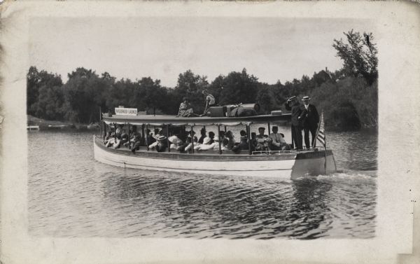 A tour boat with many passengers is traveling down a river. Two boys are sitting on the roof with suitcases and other baggage, and two men wearing hats are standing at the back near a flag. A sign on top of the boat reads: "Passenger Launch". A tree-lined shoreline is in the background.