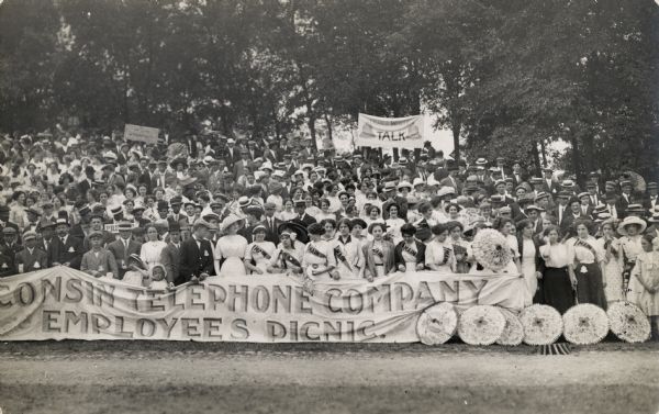 The Wisconsin Telephone Company Employees' Picnic (written on a large banner held up by the front line of people). People in the back of the crowd are holding other signs and banners, one of which reads: "Don't Write Talk".  Many of the women have sashes reading "Main". Men, women, and children make up the crowd, and a number of umbrellas are lined up on the ground.