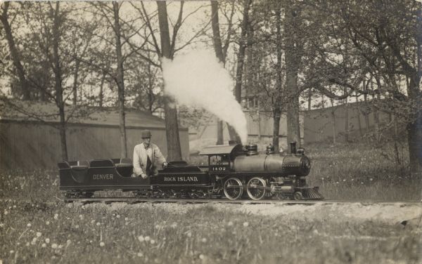 A man is sitting in the front seat of a railroad car in a small railroad train made for children, which is riding along a track in a wooded area with a building and a fence in the background.