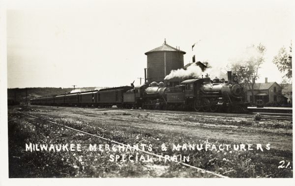 The Milwaukee Merchants & Manufacturers Special Train. A steam engine with several cars stretching to the horizon is sitting on the tracks in front of a platform and water tower. A man is standing on the first car on top of a load of coals.