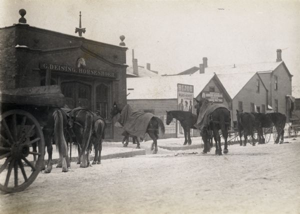 View across road towards a building with a sign on the building that reads: "G. Deising, Horseshoer". A number of horses are standing on the snowy road nearby, some covered with blankets. A man is leading one horse towards the front door.