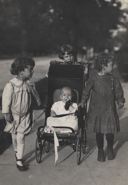 Four children are out for a walk.  One is pushing a baby (the youngest of the four) in a stroller while the other two are walking on either side.