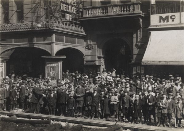 A large crowd of children are gathered outside the Alhambra theater. The children, all boys, fill the sidewalk, and are being herded by a man on the left. Standing in the arched entrances are women and girls. Posters and signs decorate the buildings.