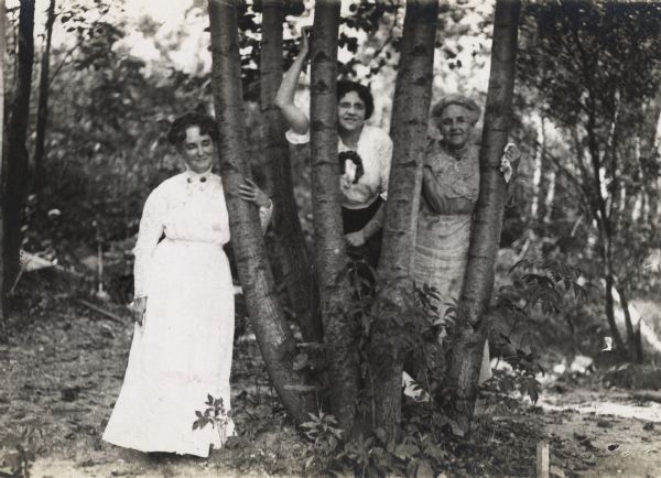 Three women are standing around and between a small set of trees in a park area. The woman in the center is identified as Alma Reinhardt Taylor; the other two are unidentified.