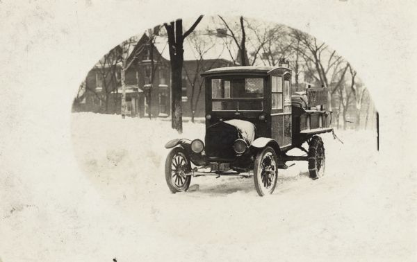 An early twentieth-century automobile parked on a road in snow, with a large house in the background. The back tires have chains around them.