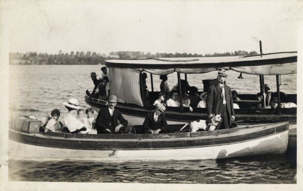 Two boats next to each other on an unidentified body of water. One boat has a roof similar to that of a tour boat. Sitting in the front boat is a family with children, with a man standing near the front smoking a pipe and holding a dog.