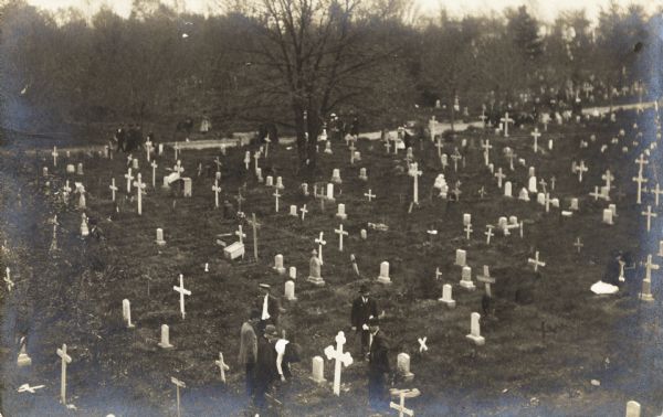Elevated view of various groups of people in a cemetery. A small group of people are gathered around a grave in the foreground. Other small groups are walking down the road in the background which is along the cemetery. On the far right, two women are kneeling before a grave.