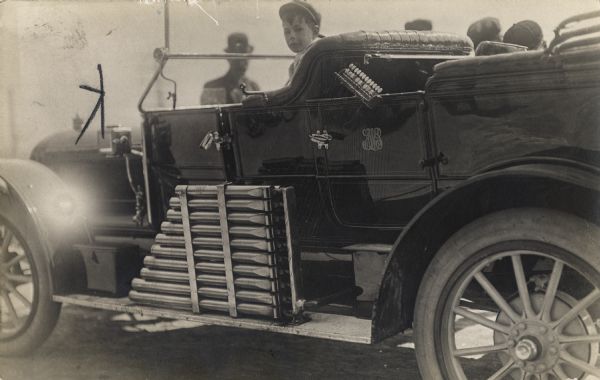 A boy in a hat is sitting in a car with an air pressure calliope attached to the side. The keyboard is attached to the driver's side rear passenger door. Men are standing behind the car, looking at the vehicle.