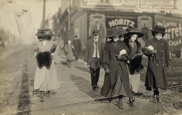 Four young women are walking down a city street across cable car tracks wearing coats and very large hats. Two of the women have fur muffs on their hands, and the other two are wearing gloves. A man is walking close behind them. Advertisements line a wall in the background.