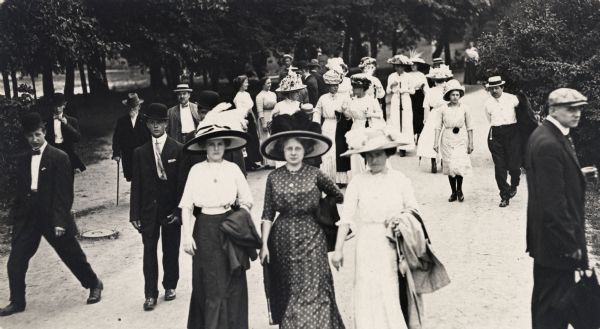 Slightly elevated view of a small crowd of well-dressed people walking down a path in a wooded park area.