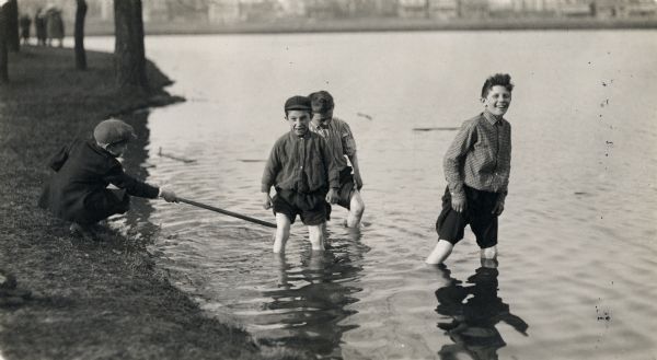 Three boys playing in the water near the shoreline with their pants rolled above their knees. A fourth boy is squatting on the grassy shore, poking one of the boys in the water with a stick.