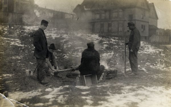 In a field with houses in the background, four men are gathered around what appears to be a campfire, from which smoke is visible. Two men are sitting, two are standing. Snow is in patches on the ground.