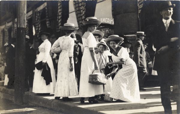 Young ladies with large straw hats standing on the sidewalk. One woman is holding a basket, while another woman is crouching down holding a purse. A child is standing with the group. Boys and men are in the background.