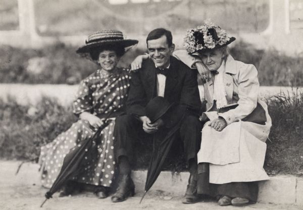 A young man and two women are sitting on a curb. Both women are wearing large hats.
