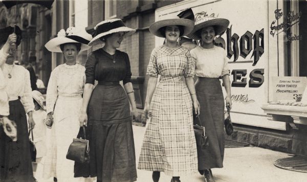 Young women carrying purses and wearing very large hats are walking down a sidewalk. A person on the left is holding an umbrella, and one woman is holding a child's hand who is walking in the background. A sign in a sweet shop window on the right advertises: "Special To-day Fresh Toasted Marshmallows".