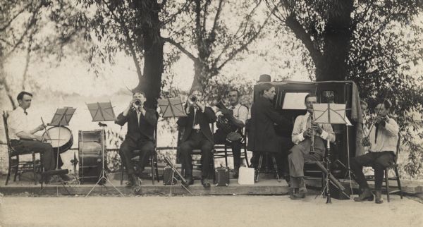 A band concert in a park. From left to right is a drummer, two trumpeters, a tuba player, pianist, oboeist, and a violinist. A boy is peeking out from behind the piano. Most of the performers are on a raised platform.