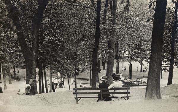 View looking down hill towards two ladies sitting on a park bench. They are looking down the hill which is surrounded by trees, watching children playing near a tree, and other people near a fence at the bottom of the hill.