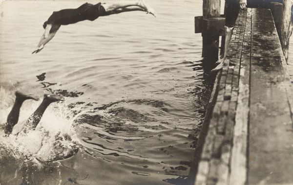 People diving from a pier into the water. On the right, a third diver is standing on the wooden pier.
