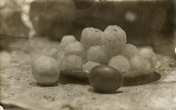 Round hailstones piled on a saucer with either grape or cherry tomatoes, or perhaps eggs, placed next to them for size comparison. A newspaper is under the items.