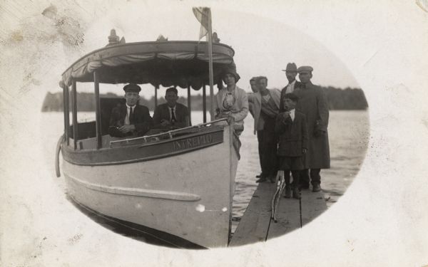 A boat named "Intrepid" near a small pier. Three people (two men and a woman), are on the boat, and five people (four men and a boy), are standing on the wooden pier.