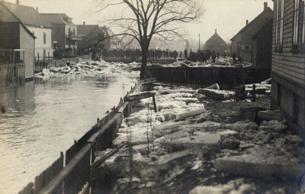 Blocks of snow and ice are piled up in a river at a bridge. A large group of people are standing on the bridge in the background. A person on the left has a long pole, trying to move some of the ice. The river is lined with houses.