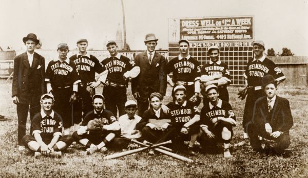 A group portrait of the Steinborn Studio baseball team, sitting and standing in a group in the outfield. The coaches are wearing suits, and the bat boys are sitting in the center. An advertisement is on the scoreboard in the background, as well as on the fence on the left.