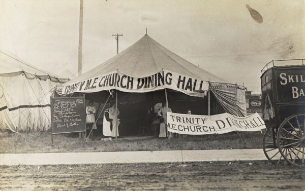 The Trinity M.E. Church Dining Hall set in a tent in a field near other tents. Two banners advertise the dining hall, and a chalkboard on the right lists the menu. A number of women are standing under the tent. An advertisement for Coca-Cola is in the background. There is a carriage in the right foreground.