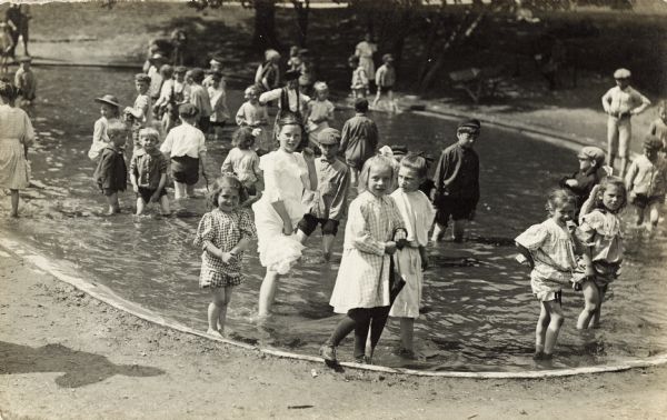 Large group of children wading in the water in a small pool. The girls are holding up the edges of their skirts, and the boys are pulling up their pants. A girl with stockings and shoes is standing on the edge of the water holding an umbrella.
