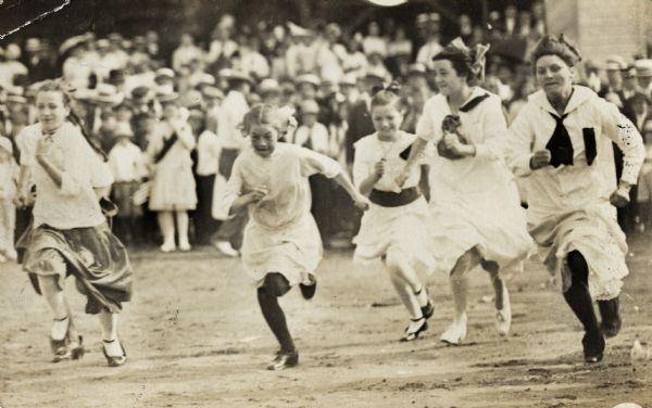 Five young girls in skirts and dresses running. In the background, a crowd is watching the race.