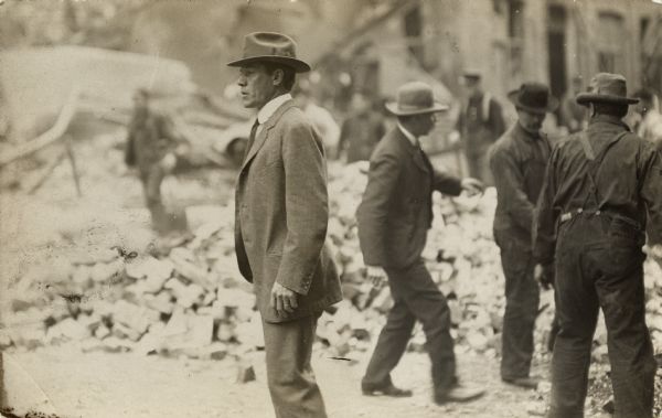 Three men are standing on the right in front of a pile of rubble. In the center, a man wearing a hat and suit is looking to the left.
