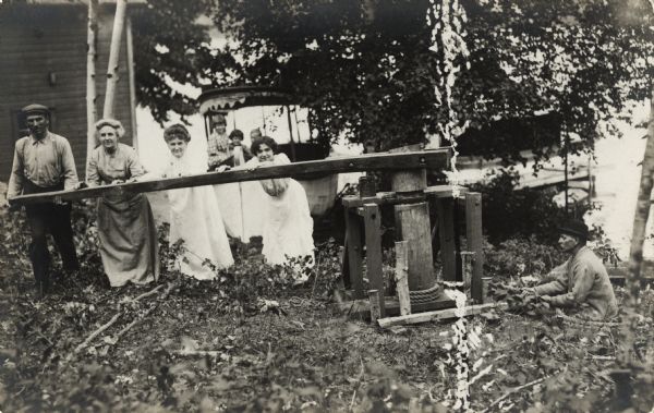 Several people next to a building in a wooded area. Four people are pushing a large wooden bar on a piece of equipment with a rope. Perhaps they are pulling up the boat from the lake. The fourth person from the left (the third woman) is identified as Alma Reinhardt Taylor, the photographer J. Robert Taylor's wife. Sitting on a boat in the background are J. Robert Taylor's children, Ellen, Donna, and Fred.