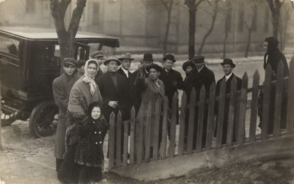 A group of fourteen people dressed for cold weather and wearing hats and scarves are standing on a sidewalk in front of a picket fence. A car is parked on the road behind them.