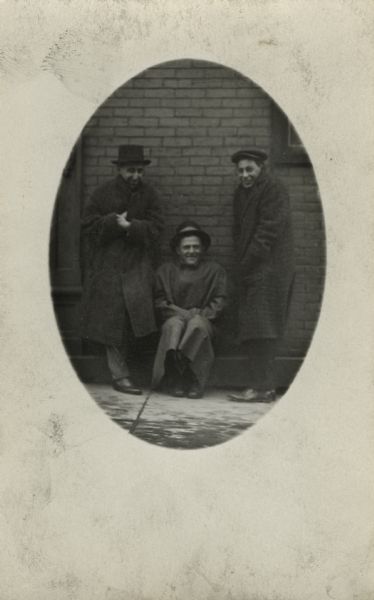 Three men wearing hats are posing against a wall, hunched up in their coats against the cold.  One stand is sitting while the other two men are standing.
