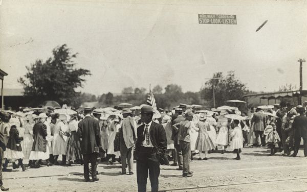 A large crowd of people ARE moving down a road. Many people are carrying umbrellas, and a flag is in the background. A sign hanging above the crowd reads: "Railway Crossing: Stop-Look-Listen".