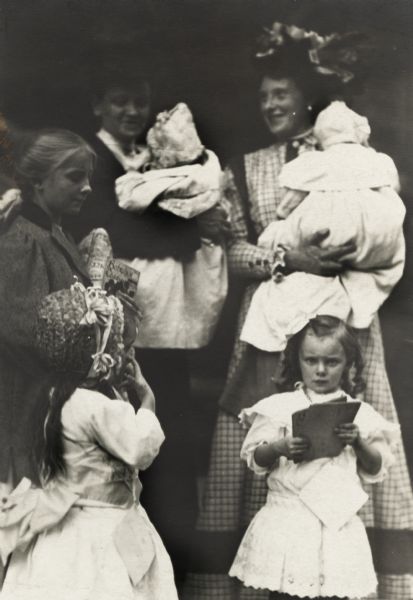 Two women wearing hats are standing and holding infants in their arms. In front of them are three girls. The tallest girl is holding some books or pamphlets, and another girl is wearing a dress and straw bonnet. The smallest girl is looking at the camera and is holding small books.