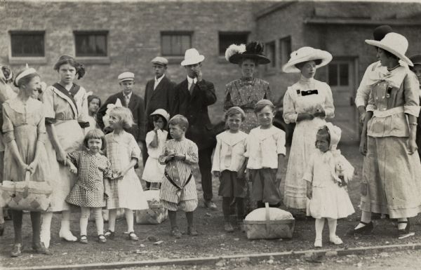 A crowd of men, women, and children gathered in front of a building with picnic baskets.
