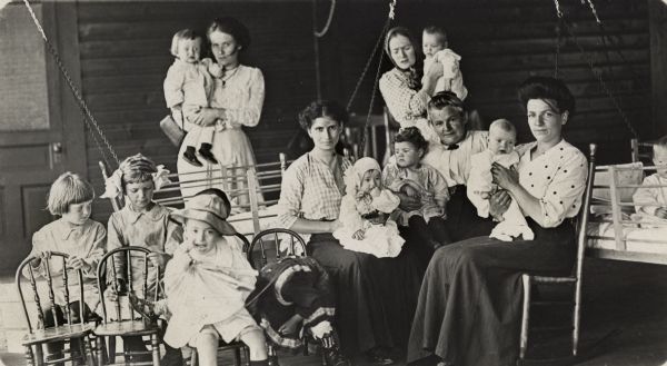 Women of various ages are surrounded by babies and young children, as well as chairs and cribs. Each woman is holding a baby. Cribs are hanging on chains from the ceiling.