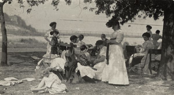 A group of women and girls and one man are sitting outside on benches and chairs sewing clothes and talking. On the right, a woman has a baby stroller beside her. In the background is a shoreline and water.
