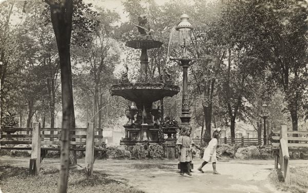 Two young girls playing on a pathway near a large fountain and lamppost located in a park.