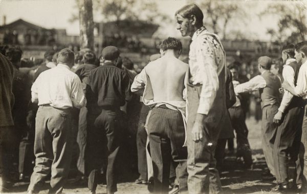 A group of men with their backs to the viewer are gathering in a circle around an unknown event. One man near the center has his shirt open in the back. In the foreground is a tall man standing in profile with dirt on his face, neck, shoulders, and back.