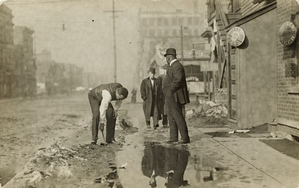 Men standing around a puddle alongside a road near a small snowbank. One man is bending down near the puddle with a hammer in his hand. Signs for beer are on the building to the right.