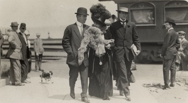 Two men escorting a fashionably dressed woman with a feathered boa obscuring part of her face. People are standing in the background near a train next to the platform. A dog is running on the sidewalk on the left.