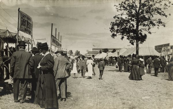People strolling on the midway and stopping to observe the booths and displays. In the background is a large building with flags.