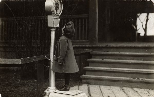A young girl wearing a coat and hat is standing on a scale that is chained to a post near a building.