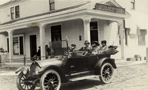 Three people sitting in the back seat of a parked car drinking beer and other beverages in glasses. The building behind them is the Otto Sprenger Millersville House, which features a sign for beer over a front porch. Two people are sitting on the porch with two young children.