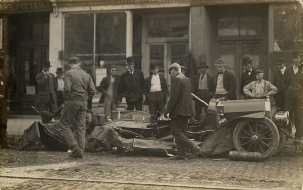 Crowd of men gathered around a car at the curb. The back of the car appears almost crushed while portions of the front remain relatively intact. People are standing on the sidewalk behind the car.