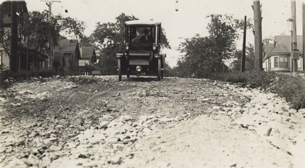 A man is driving an automobile through a neighborhood on a dirt and rock road.