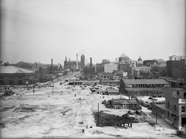 Elevated view of industrial area. The building in the foreground has a sign that reads: "Machine Shop" on it roof. People are working in a large open area, and in the background is part of the downtown, with streets and a building with a clock tower.