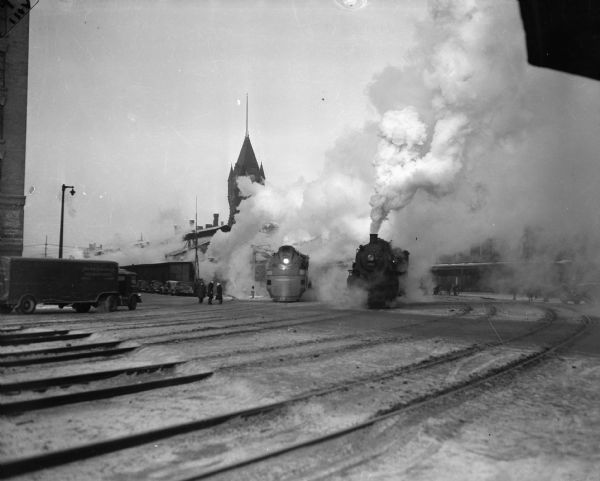 View across multiple sets of railroad tracks towards two locomotives at a railroad city yard in Milwaukee (probably the Hiawatha). There is a dense cloud of steam surrounding them, and three men are standing on the left side.